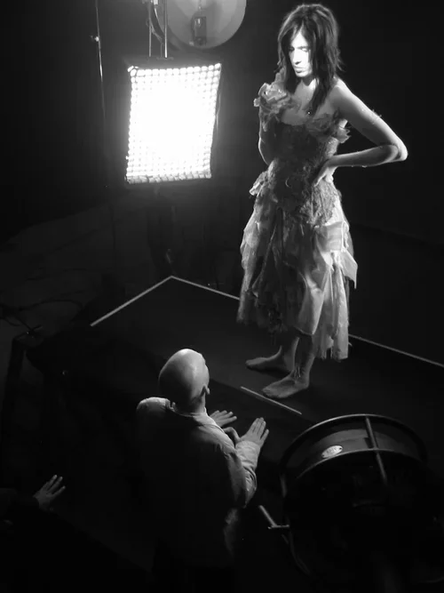 A woman in an elaborate dress stands on a platform in front of a bright light, with her hand on her waist. A person in the foreground looks up at her, with a camera or light stand nearby.