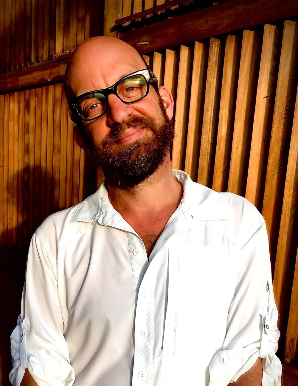 A man with a beard, glasses, and a white shirt standing against a wooden background with vertical slats.