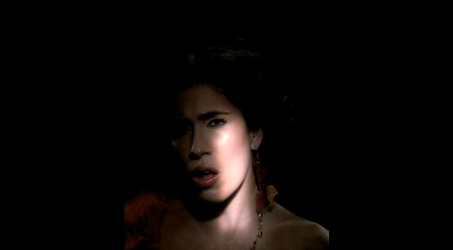 A woman with dark hair, wearing makeup, with a serious expression, illuminated in a dark environment.
