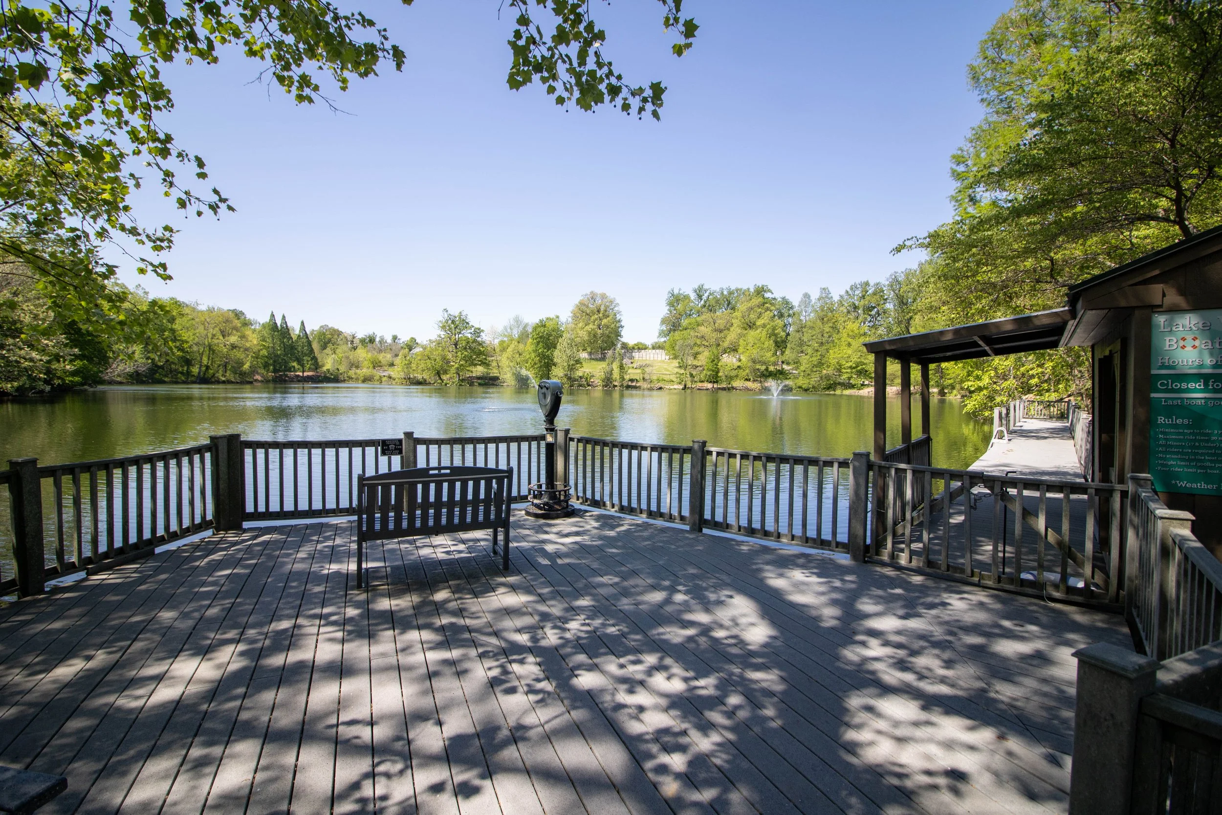 A wooden deck by a lake with a railing, a bench, and a sign, surrounded by green trees under a clear blue sky.