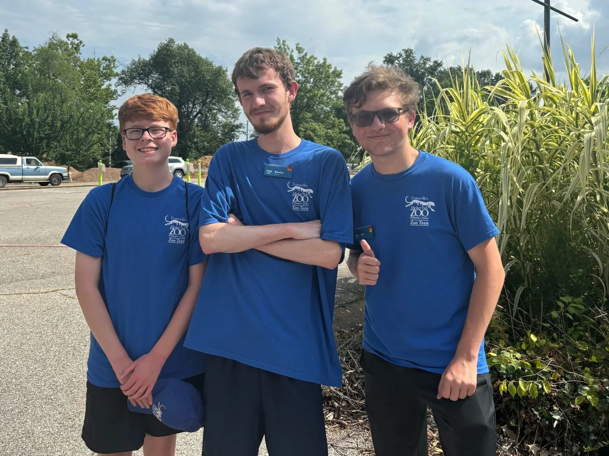 Three young men with blue shirts standing together in front of some plants with vehicles in the background.