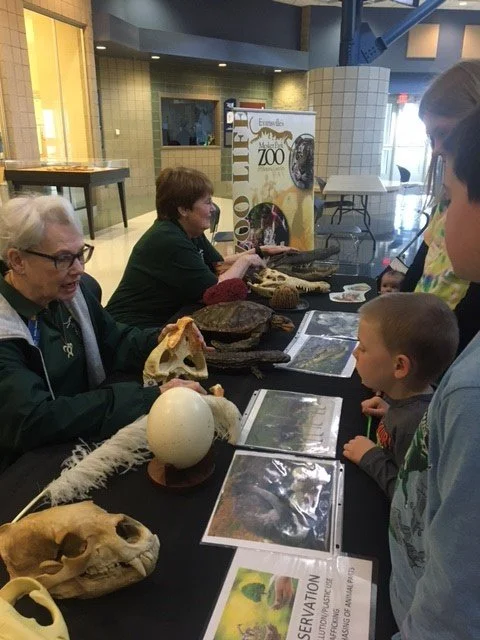 Two zoo volunteers in green shirts at a table with guests. Table is covered with various animal biofacts like skulls, feathers, and eggs.