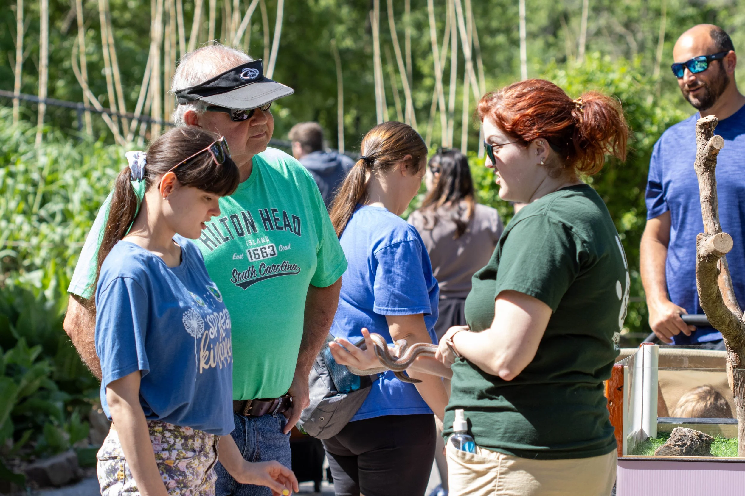 People standing outdoors in a queue, some wearing sunglasses, engaging in conversation with a woman holding a snake. Bright sunlight, green trees, and a natural setting.