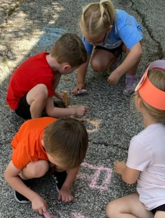 Four children drawing on the pavement with chalk, one girl wearing a pink headband and white shirt, one girl in a blue shirt, one boy in a red shirt, and another boy in an orange shirt.