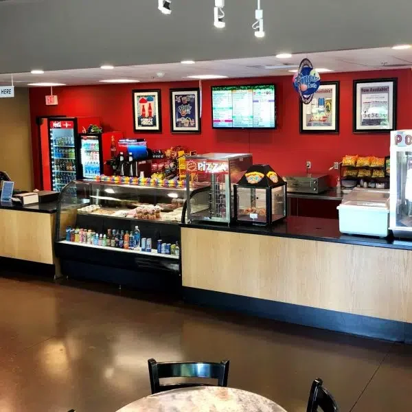 Inside look at a pizza restaurant's counter with a variety of snacks and drinks, a digital menu board, and red walls decorated with framed posters.