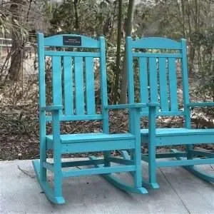 Two blue wooden rocking chairs outdoors on a wooden deck with trees and bushes in the background.