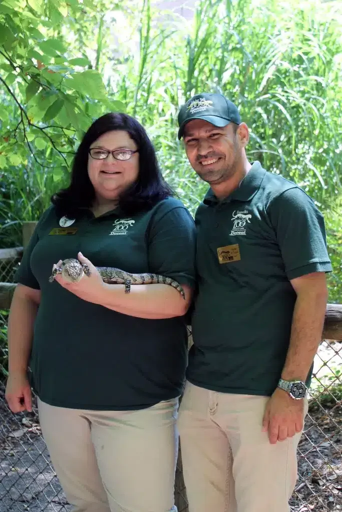 Two zoo employees in green shirts and beige pants standing outdoors among greenery, holding a small alligator or crocodile.