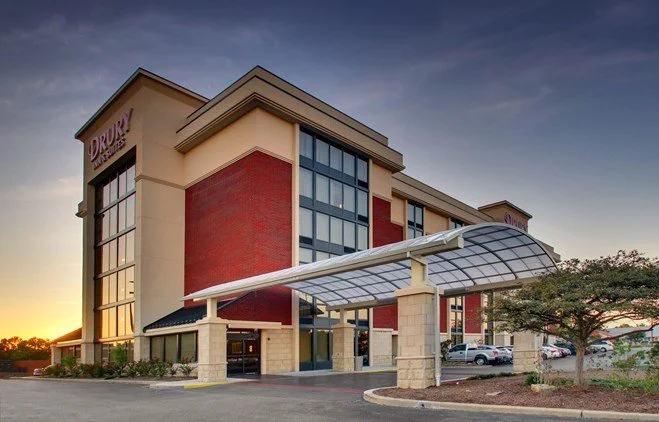 Mid-rise modern building with a red brick exterior and large windows, featuring a covered entrance canopy, parking lot, and trees in the foreground.