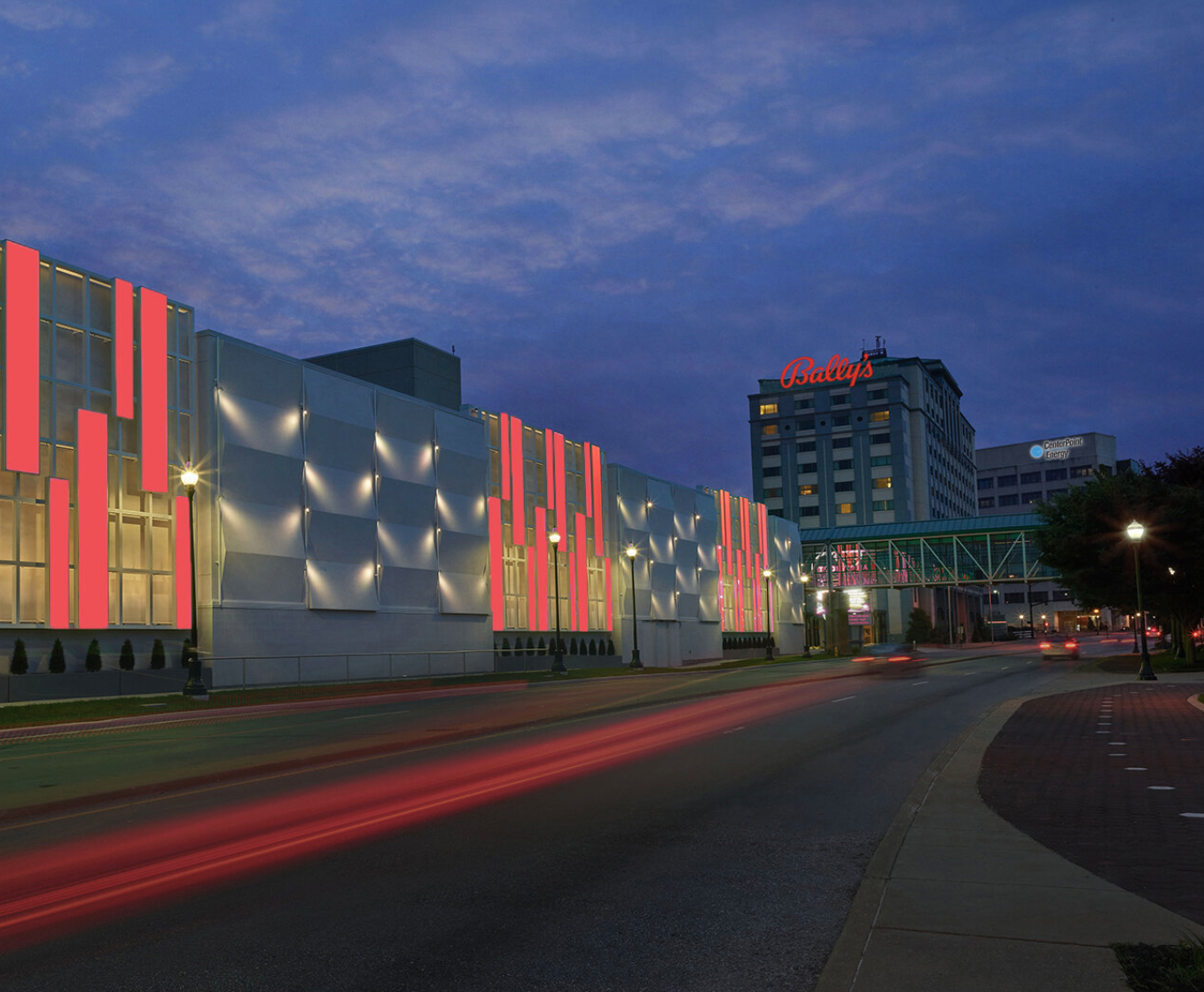 City street at dusk with illuminated modern building featuring pink and white accents, and taller buildings in the background, including one with a red 'Bally's' sign.