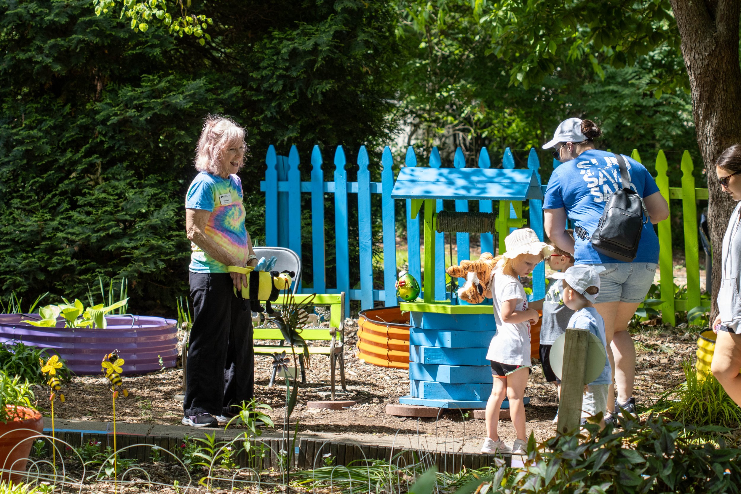 A group of people, including children and adults, gathered outdoors in a lush garden with a blue and green fence, engaging in gardening or outdoor activities on a sunny day.