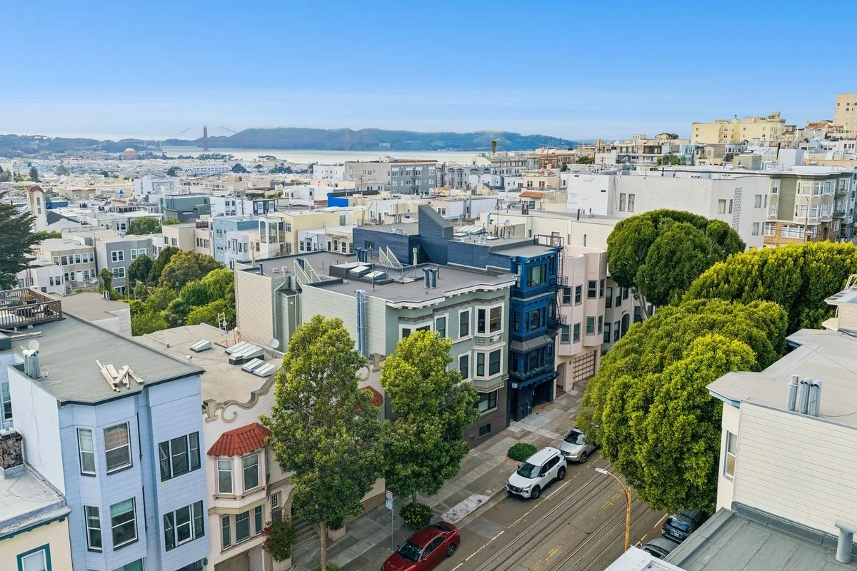 View of San Francisco neighborhood with colorful row houses, trees, parked cars, and iconic Golden Gate Bridge in the distance under a blue sky.