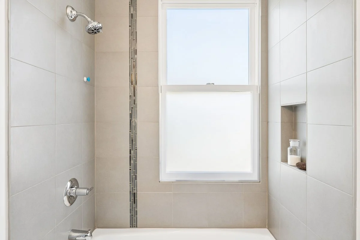 Empty shower with beige tiles, a large frosted window, and built-in shelf with soap and a jar.