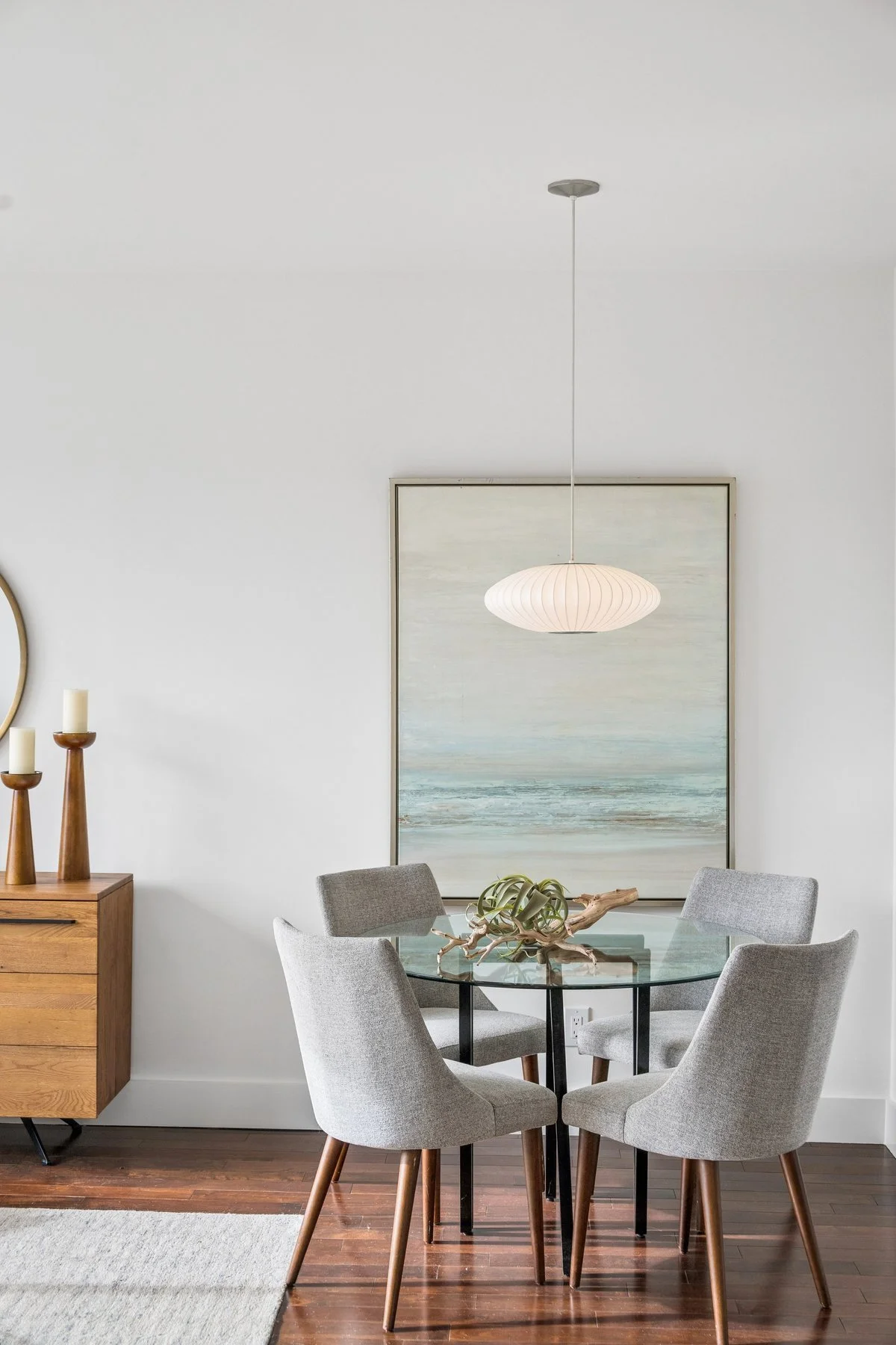 Living room dining area with a round glass table, four light gray upholstered chairs, a large abstract ocean painting, a white pendant light, and a wooden sideboard with candles.