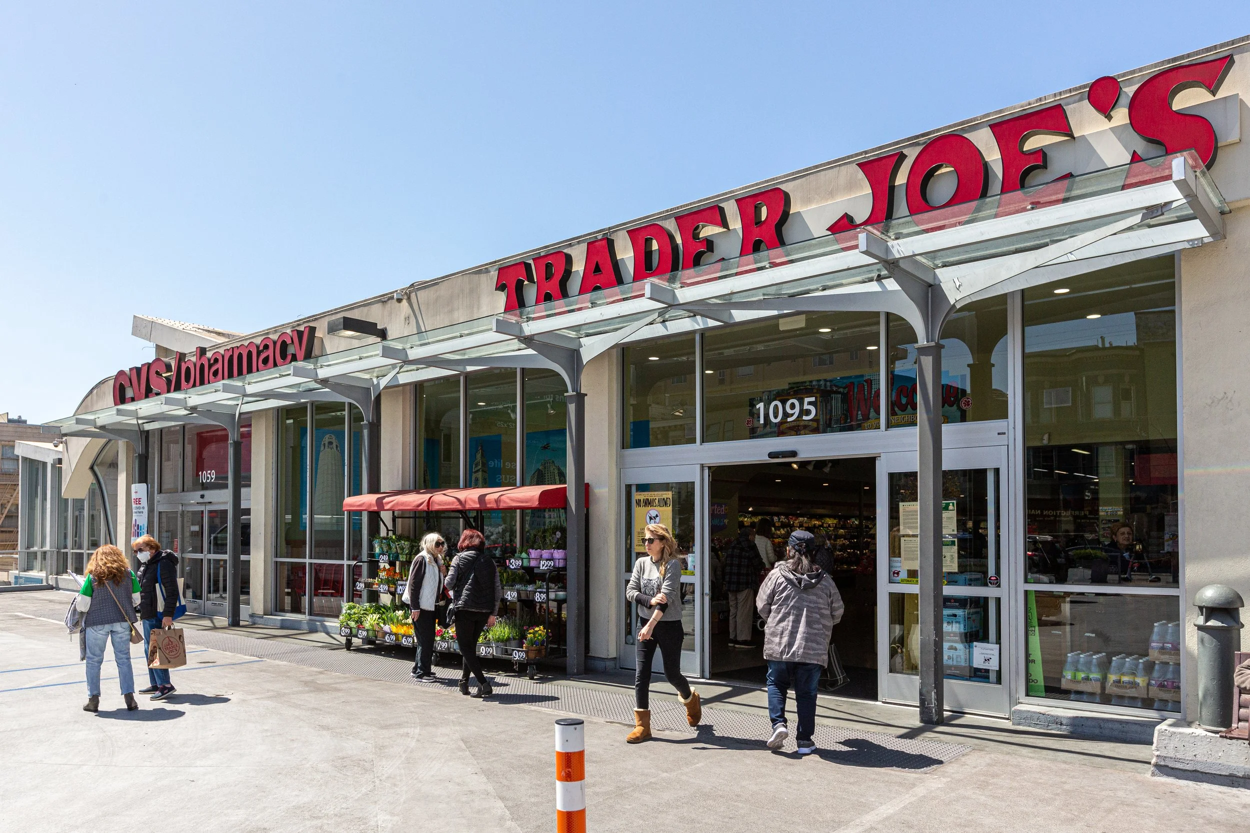 Exterior of a Trader Joe's grocery store with people entering and exiting, signage visible, and outdoor display of flowers.