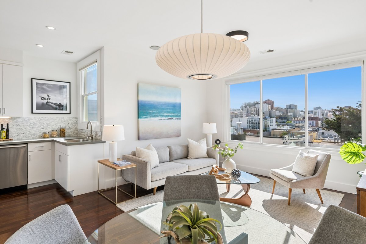 Living room with white walls, large window showing city view, modern furniture including beige sofa, armchair, coffee table, and a dining table, with artwork and plants, bright and airy.