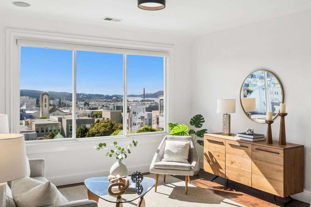 Living room with large window view of city skyline and bridge, inside decor includes a sofa, armchair, wooden sideboard with mirror, lamps, and decorative items.