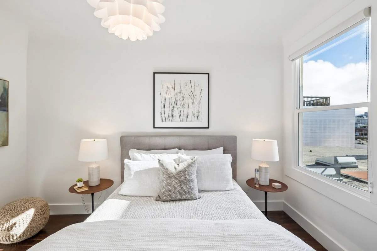 A neatly made bed with multiple white pillows and a textured gray pillow in the center, in a bright bedroom with a window showing a city view, two matching bedside tables with lamps, minimalist wall art above the headboard, and a woven pouf on the fl