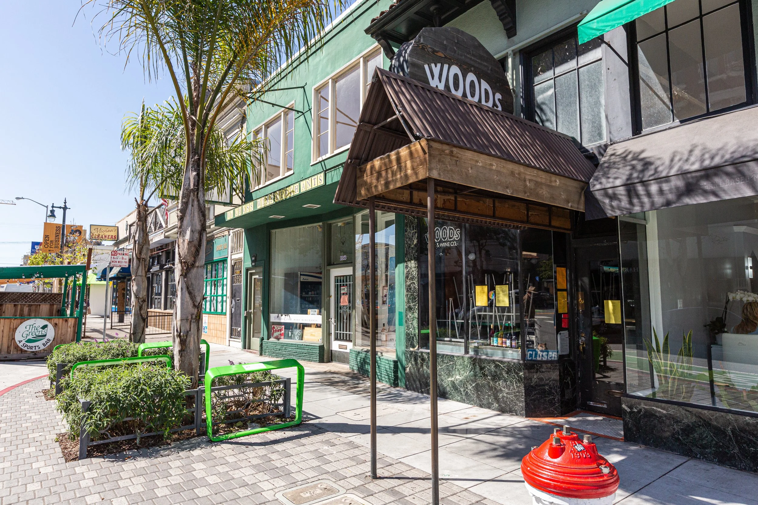 Street view of a building with a sign that says 'WOODS' and a smaller sign that reads 'GOURMET AND ANTS.' Two palm trees are in front of the building, with a sidewalk and a small garden area enclosed by a green metal fence. There is a large, round, o