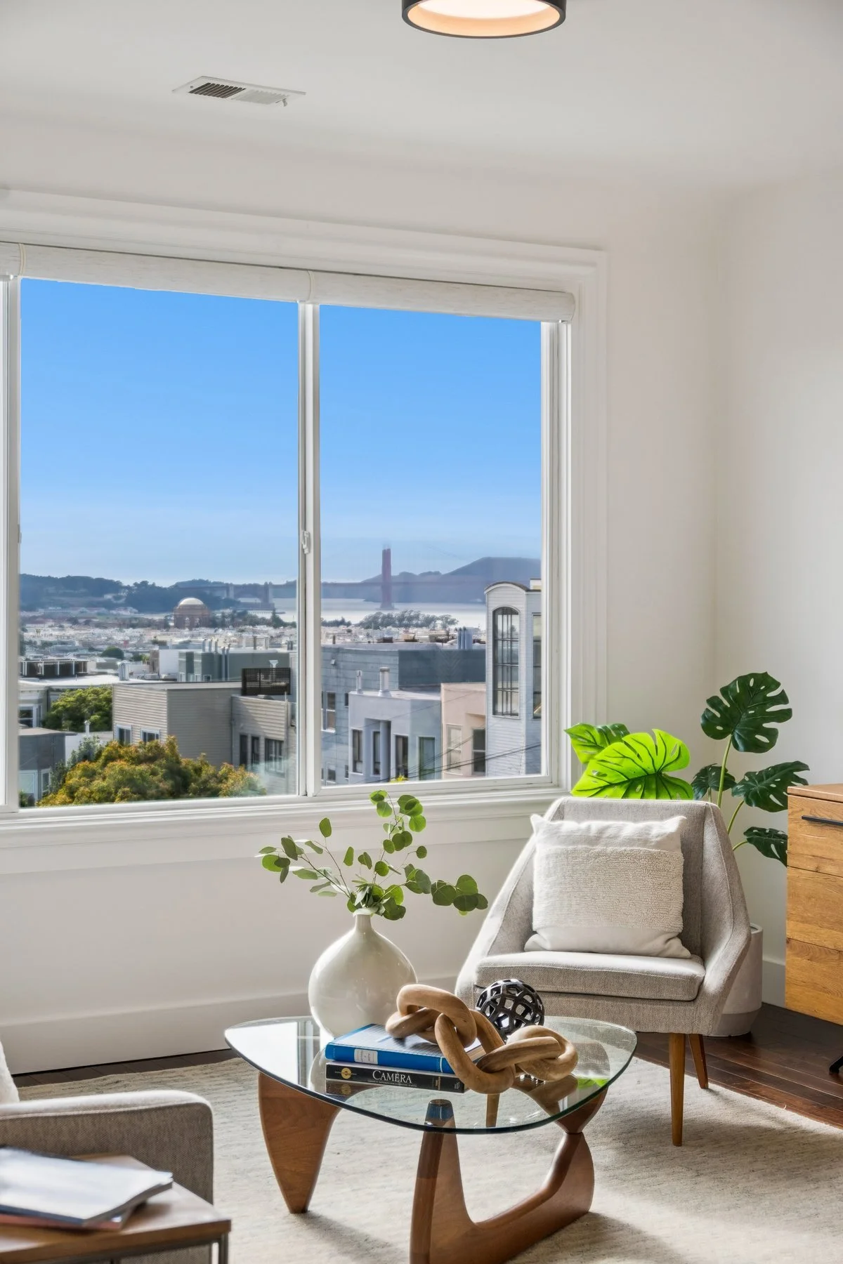 Living room with a large window showing a city view of San Francisco, including the Golden Gate Bridge. The room has white walls, a beige chair with a pillow, green houseplants, a glass coffee table with books and decorative objects, and a wooden dre