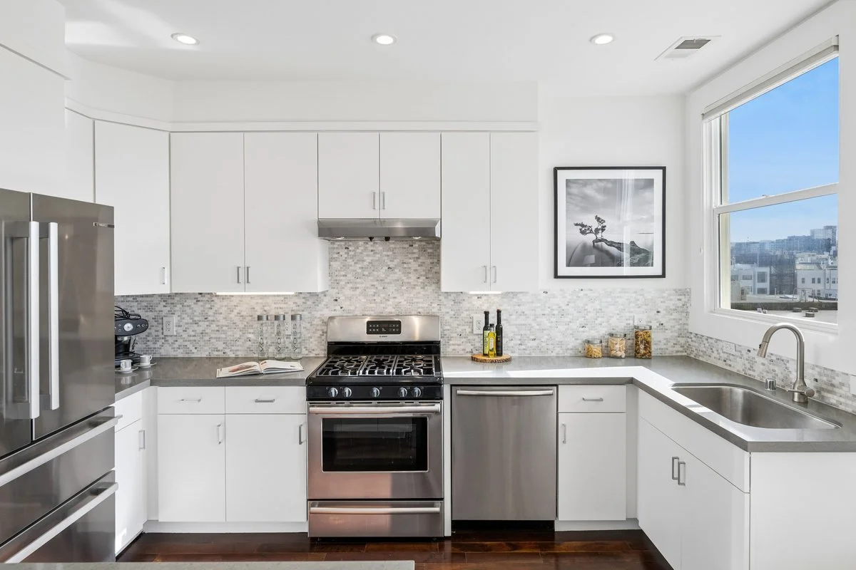 Modern kitchen with white cabinets, stainless steel refrigerator, oven, dishwasher, and a sink under a window, with a mosaic tile backsplash, decorated with a framed black and white picture on the wall.