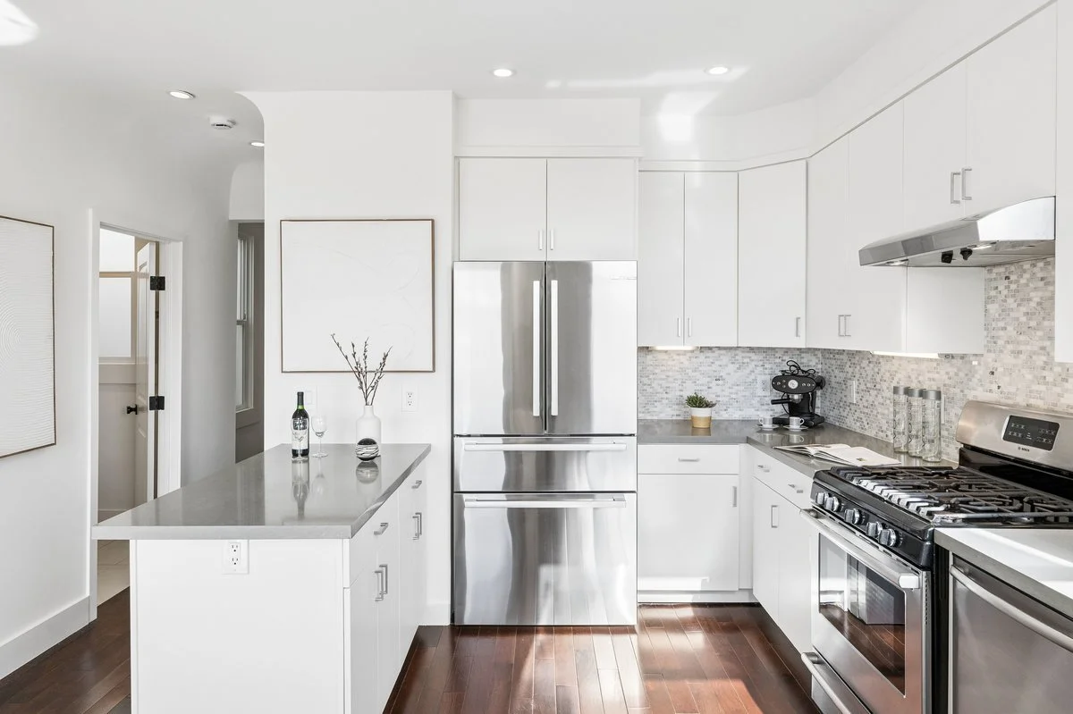 Modern white kitchen with stainless steel appliances, including a refrigerator, stove, and microwave, with a small island featuring a vase with branches and two wine glasses.