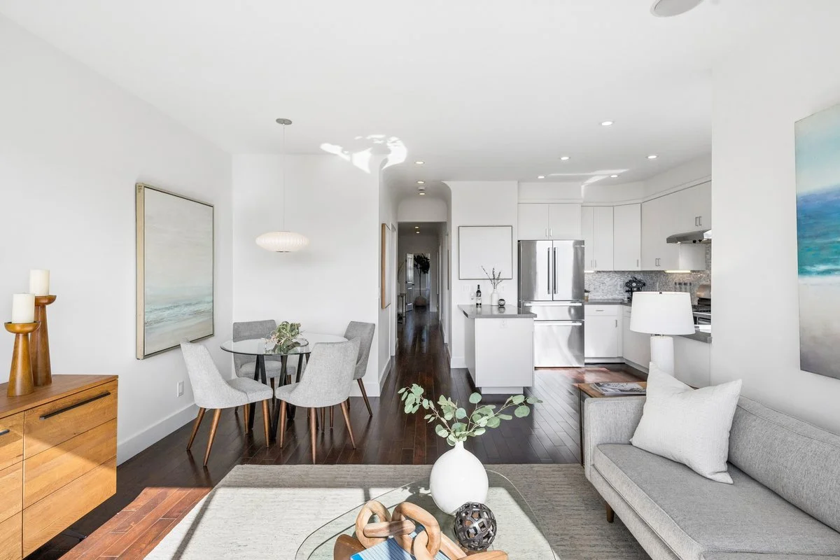 Bright and modern living room and kitchen with white walls, wooden flooring, gray sofa, dining table with chairs, and contemporary art.