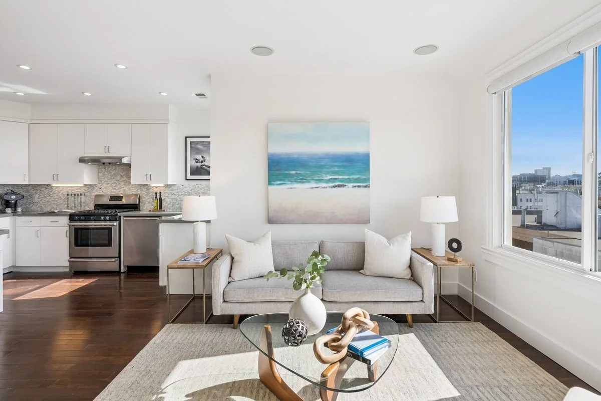Living room with a beige sofa, two side tables with lamps, a glass coffee table with decorative objects, a painting of the ocean above the sofa, and a large window showing city buildings outside. Part of the kitchen with white cabinets, stainless ste