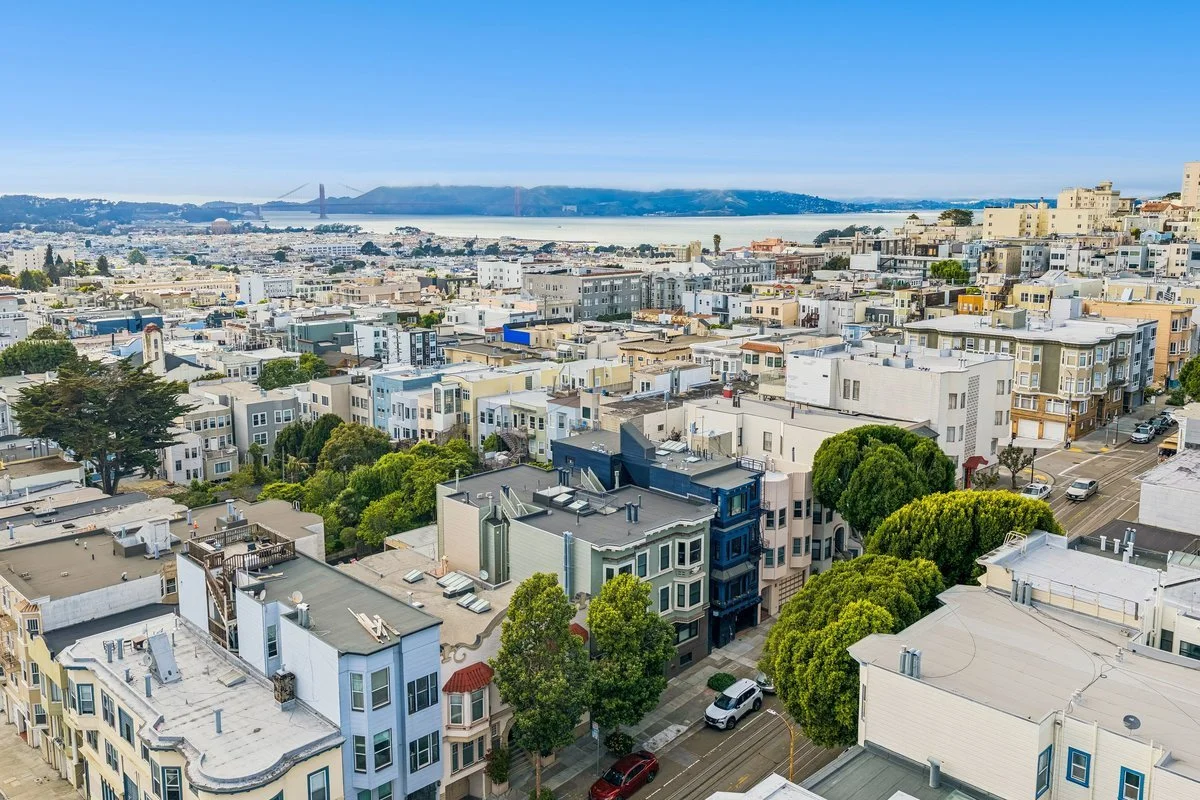 Aerial view of a city with colorful buildings, green trees, streets, and cars, with a body of water and hills in the background, under a clear blue sky.