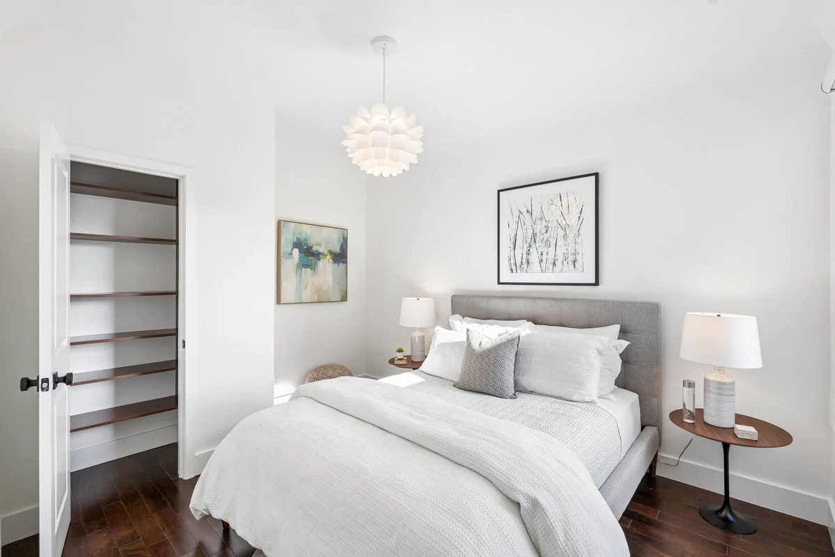 Clean, modern bedroom with white walls, gray upholstered bed, white bedding, decorative pillows, two matching bedside lamps, and minimalist artwork on the wall.