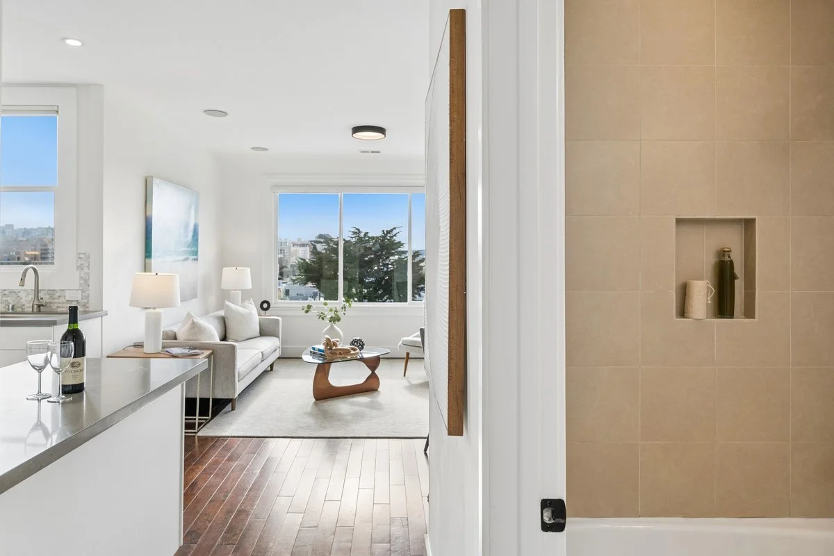 Interior view of a modern apartment with a living room and bathroom. The living room has a sofa with pillows, a coffee table, two lamps, and a large window showing trees and buildings outside. The bathroom features beige tiled walls with a small rece