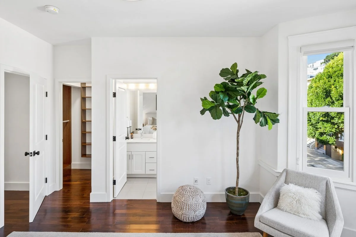 Bright, modern living space with white walls, hardwood floors, a large window with greenery outside, a potted plant, a cozy chair with a white pillow, and an open door leading to a bathroom.