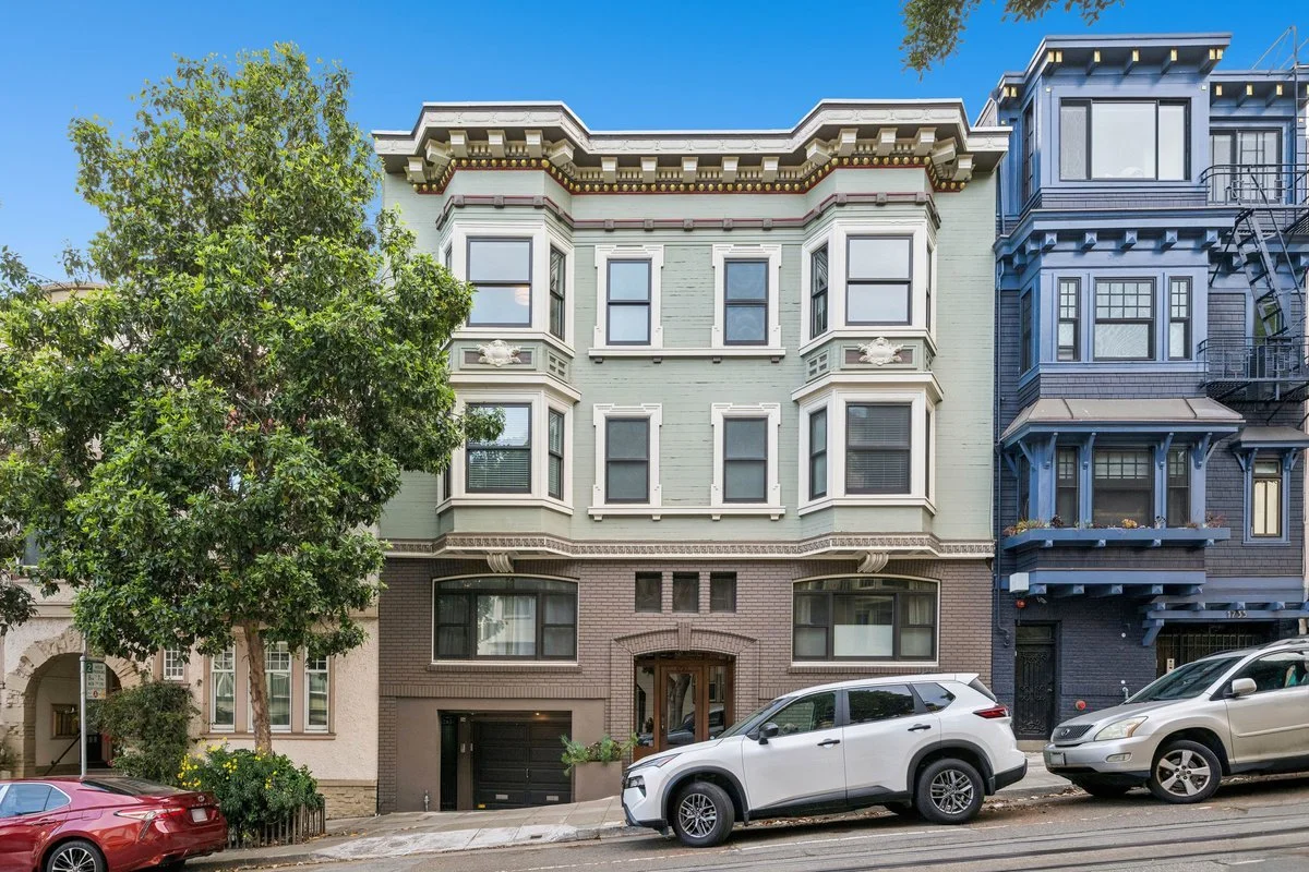 A multi-story residential building on a city street with parked cars and a tree in front.