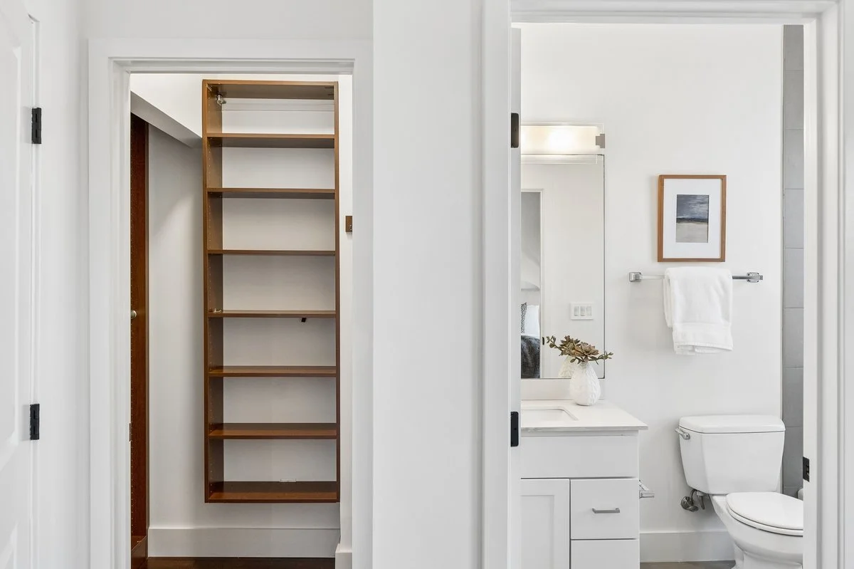 Empty closet with wooden shelves next to a bathroom with a sink, mirror, and toilet.