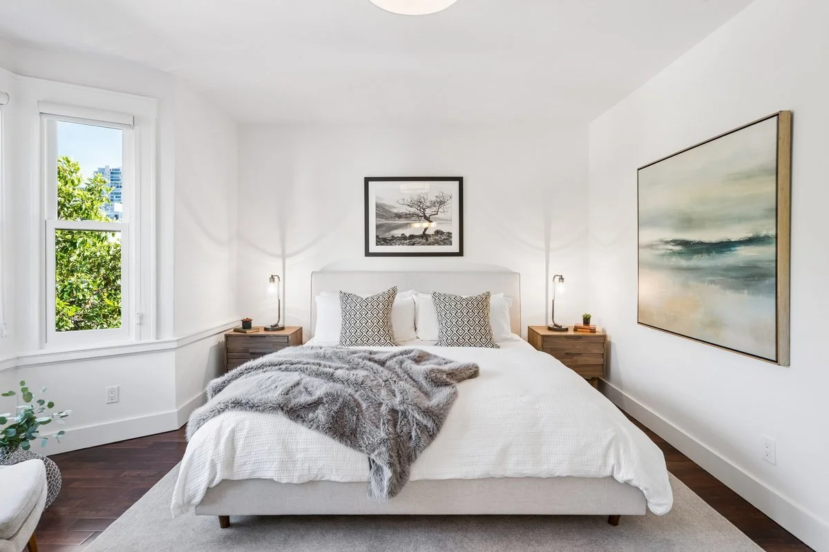 Bright bedroom with white walls, a large bed with white bedding and patterned pillows, twin nightstands with lamps, and framed artwork on the walls. A window shows greenery and city buildings outside.