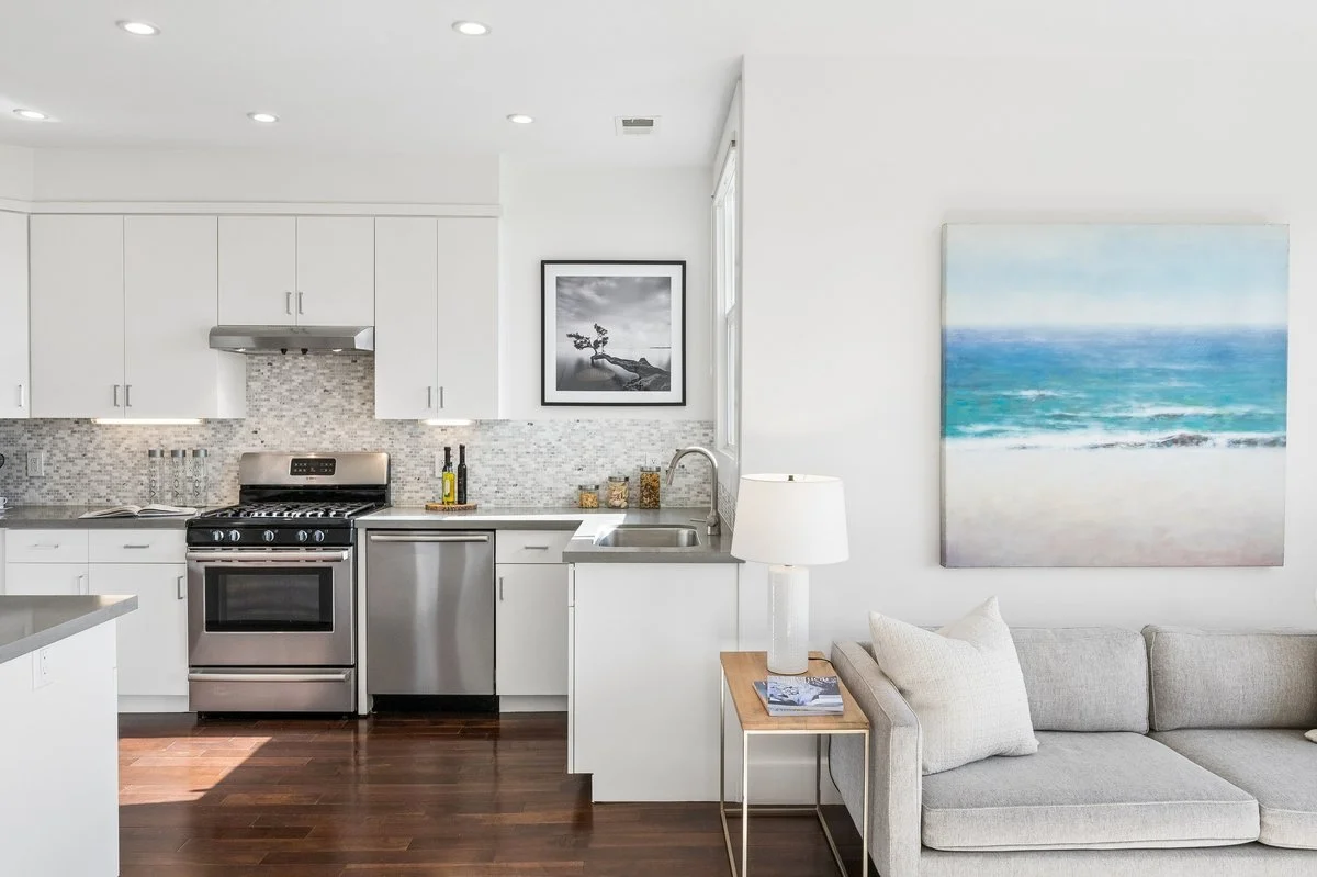 Open-concept living room and kitchen with white cabinets, stainless steel appliances, hardwood floors, a gray sofa, and ocean-themed artwork.