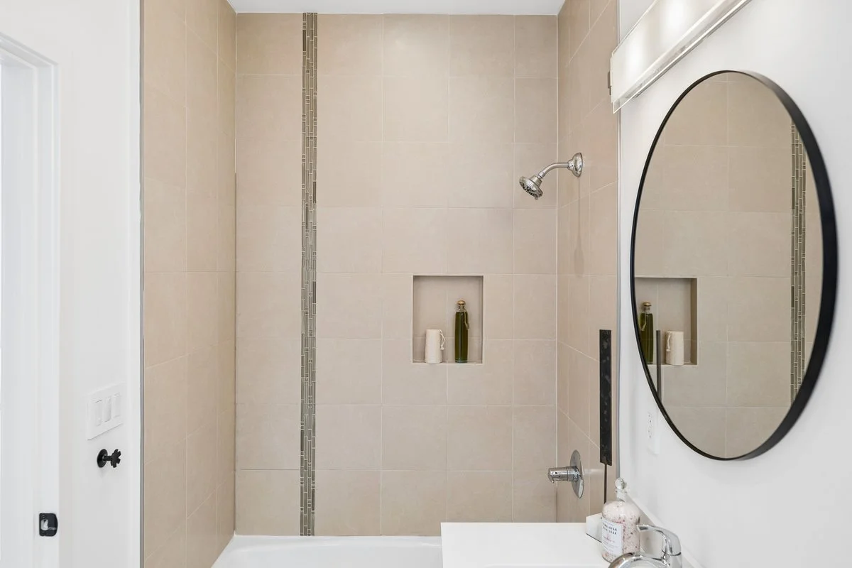 Bathroom shower with beige tile walls, decorative tile strip, niche with bottles and toiletries, round mirror, and a faucet with a soap and lotion dispenser.