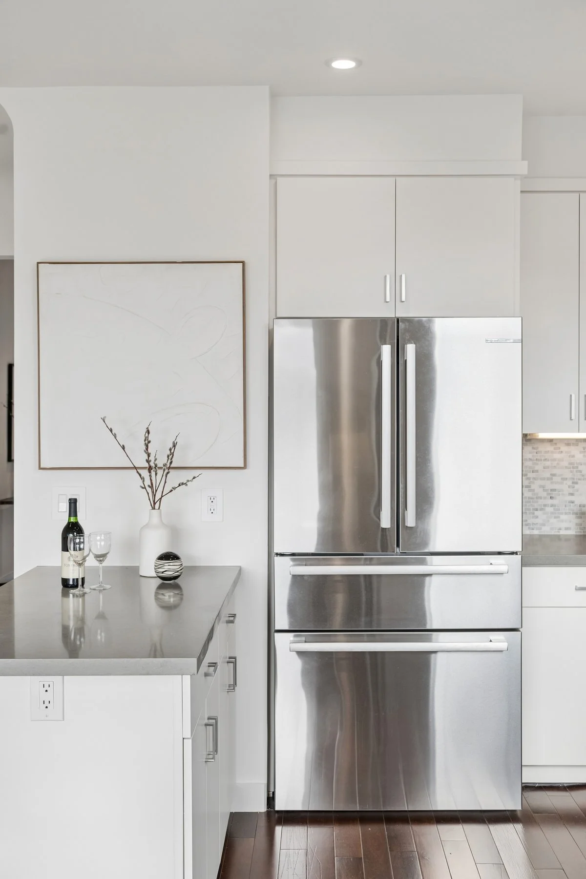 Modern kitchen with stainless steel refrigerator, white cabinets, gray countertop, and decorative items like wine bottle, glasses, and a vase with branches.