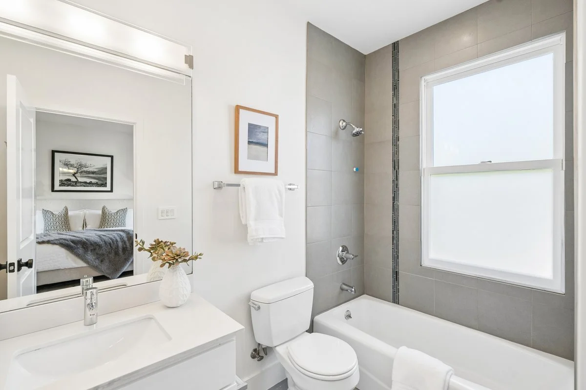 A clean, modern bathroom with a white vanity, a large mirror, a toilet, and a bathtub with a glass window letting in natural light. The bathroom has neutral-colored tiles and minimalist decor.