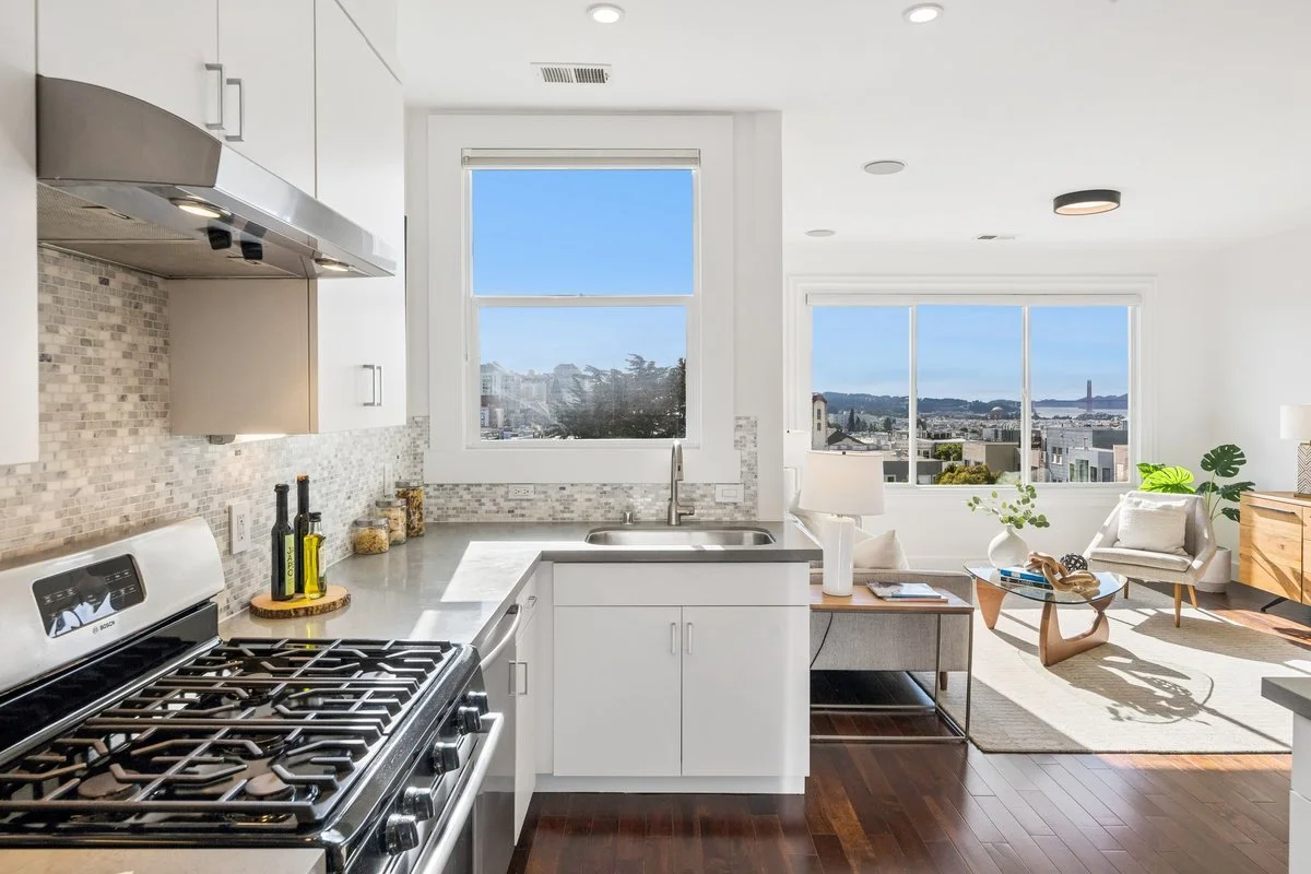 Modern kitchen with white cabinets, stainless steel appliances, beige mosaic backsplash, gray countertops, and a view of a living room and windows with city and water views