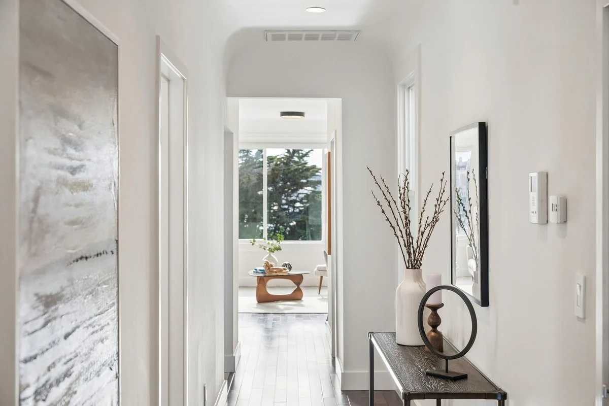 Bright hallway leading to a sunlit living space with large window, wooden table, and plants.