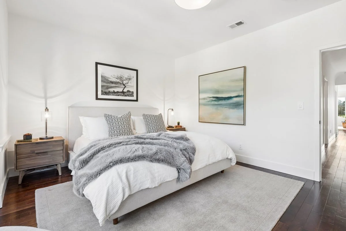 Modern bedroom with white walls, a bed with white bedding, and gray throw blanket. Two nightstands with lamps and decorative items. Wall art above the bed and on the adjacent wall. Hardwood flooring with a light gray rug.