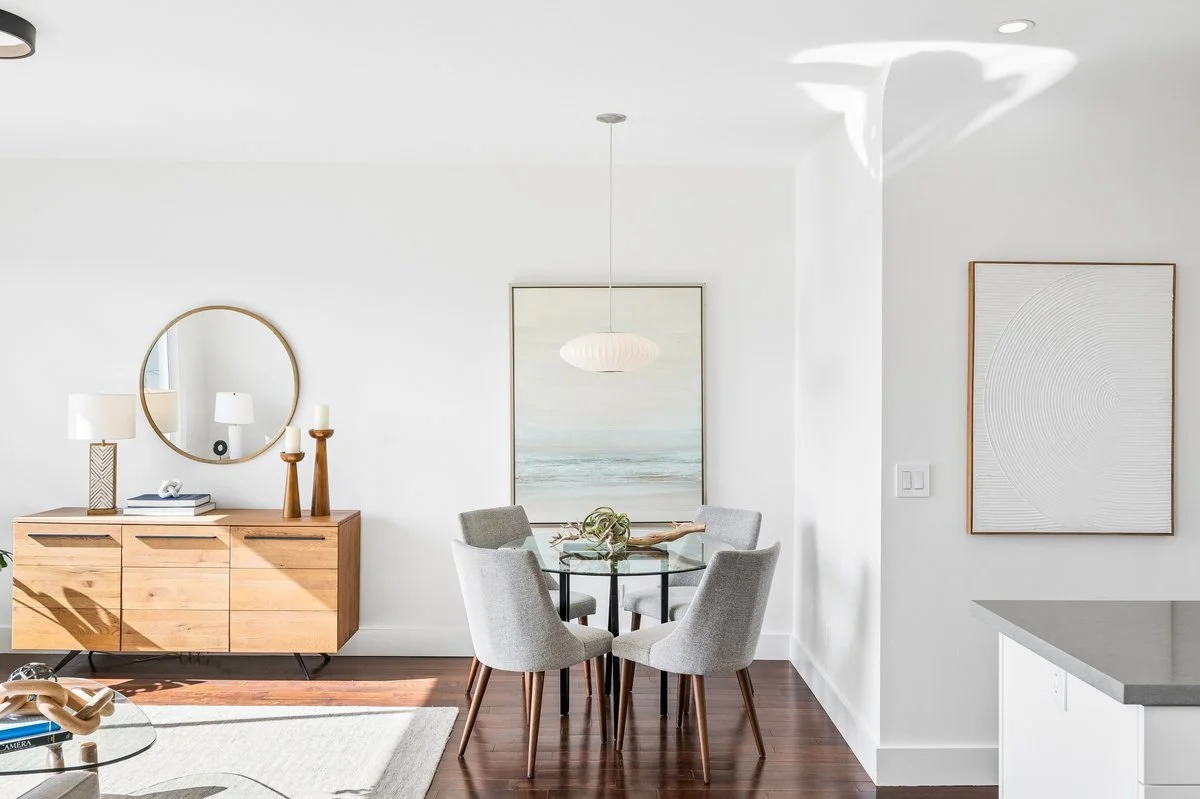 Modern dining room with a round glass table surrounded by five gray upholstered chairs, a wooden sideboard with decorative lamps and candlesticks, a round mirror, abstract wall art, and a paper pendant light, with hardwood floors and light-colored wa