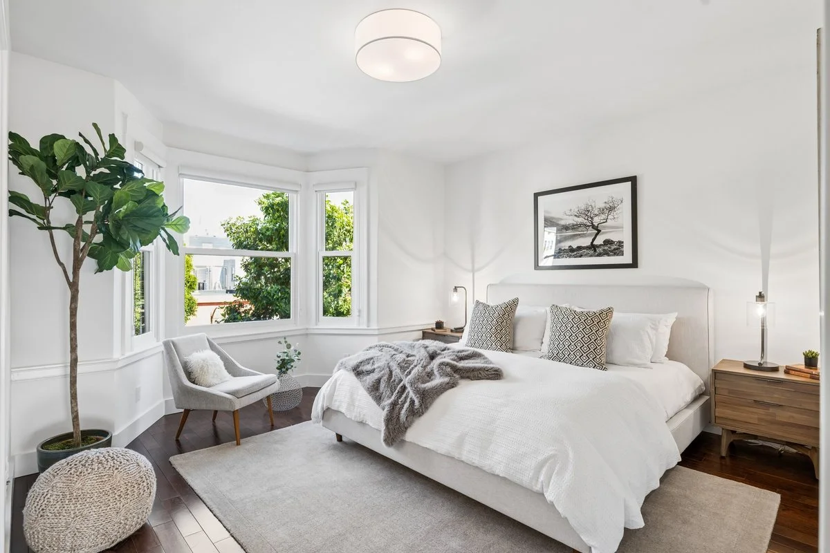 Bright bedroom with white walls, a large bed with white bedding and decorative pillows, a wooden nightstand with a lamp, a chair with a pillow, a potted plant, and a framed black-and-white nature photo on the wall.