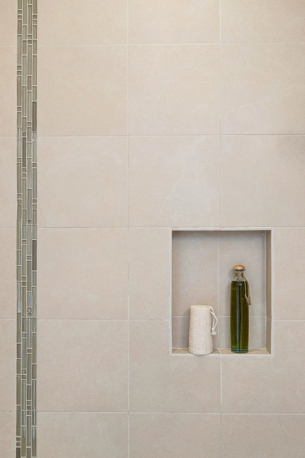 A built-in wall niche in a tiled bathroom wall holding a small beige bottle and a tall green glass bottle.