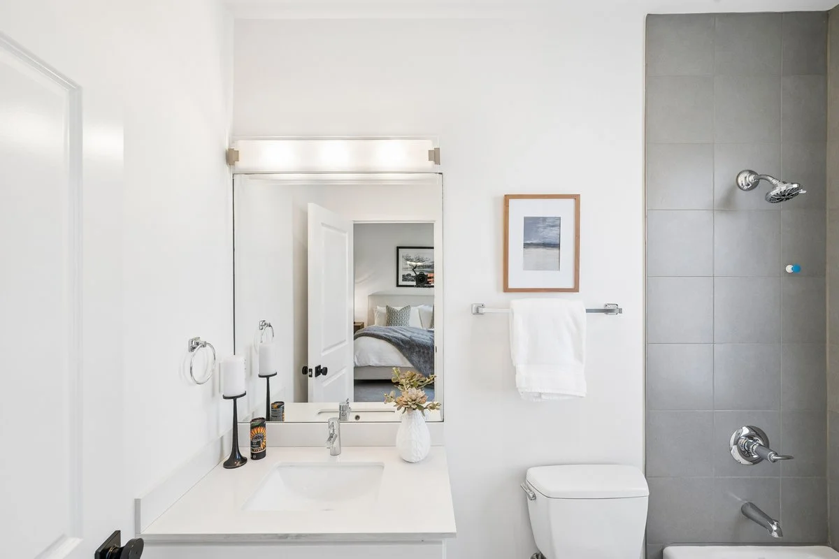 A clean and modern bathroom with white walls, a sink with a mirror, a framed picture, a towel rack with a white towel, a toilet, and a shower area with gray tiles and silver fixtures.