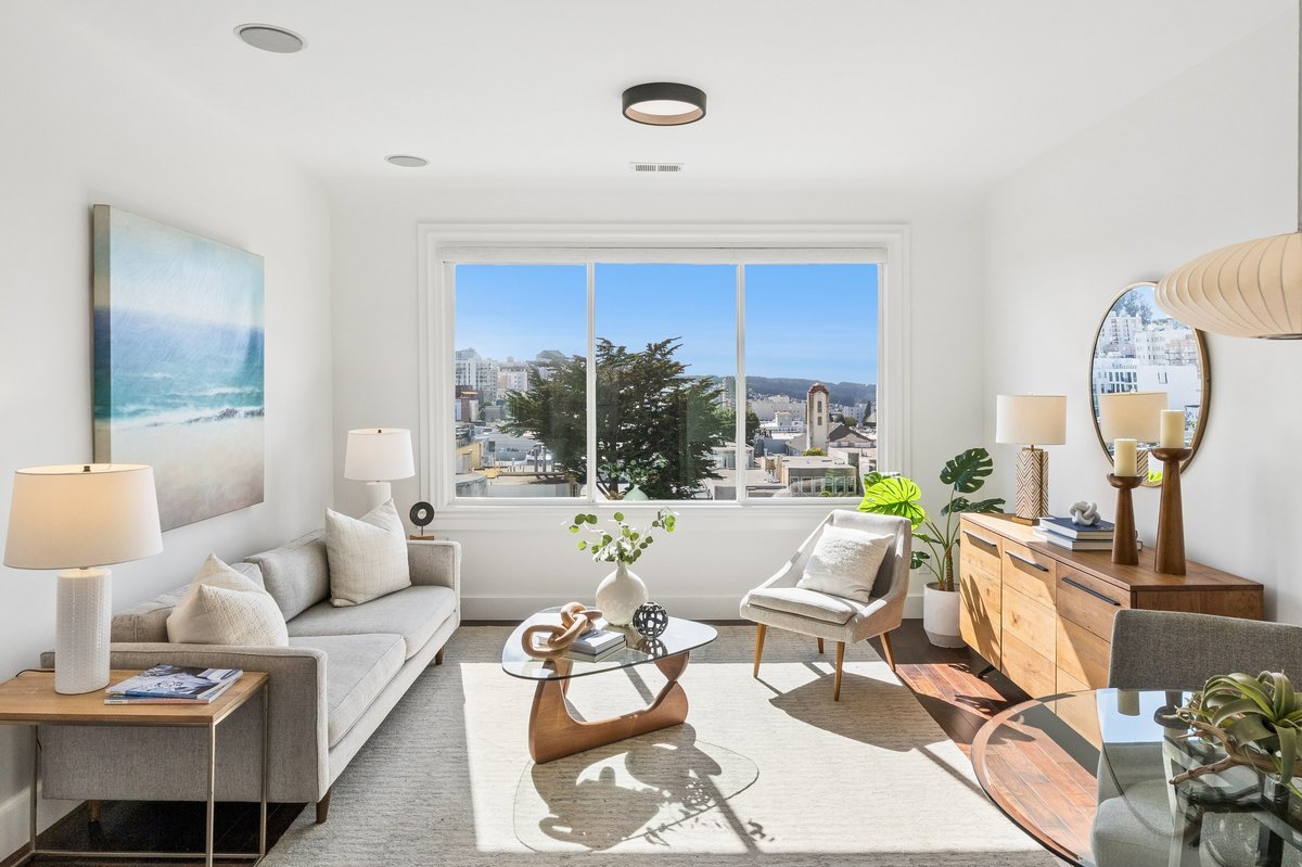 Bright living room with large window showing cityscape, featuring a light gray sofa, side tables with lamps, a coffee table with decorative items, a white armchair, a wooden sideboard with lamps, and a mirror reflecting part of the room.