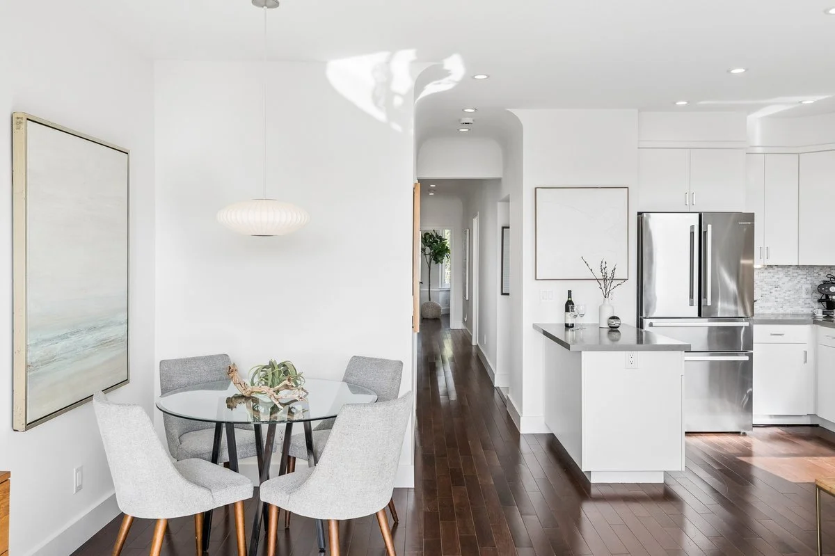Modern kitchen and dining area with white walls, dark hardwood floors, a round glass table with four gray upholstered chairs, a pendant light, and stainless steel appliances.