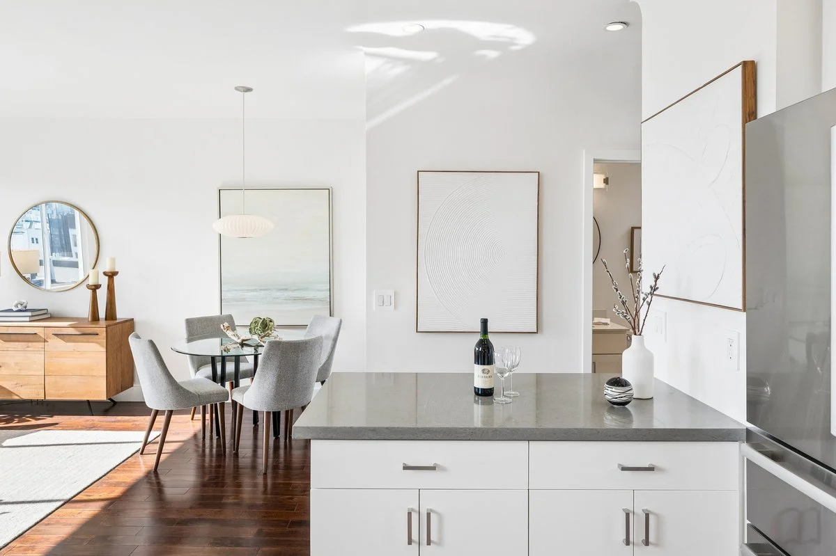 Modern open-concept living space with a kitchen and dining area, featuring white walls, dark wooden floors, a round glass dining table with gray chairs, a wooden sideboard with decorative items, and abstract wall art.