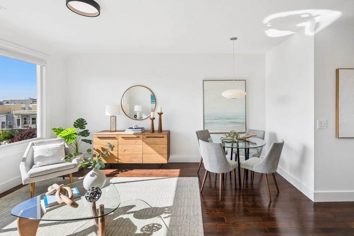 Bright dining area with a glass-top round table and four gray upholstered chairs, a wooden sideboard with a mirror above, a white armchair with a cushion, and large window with a view of neighboring buildings.