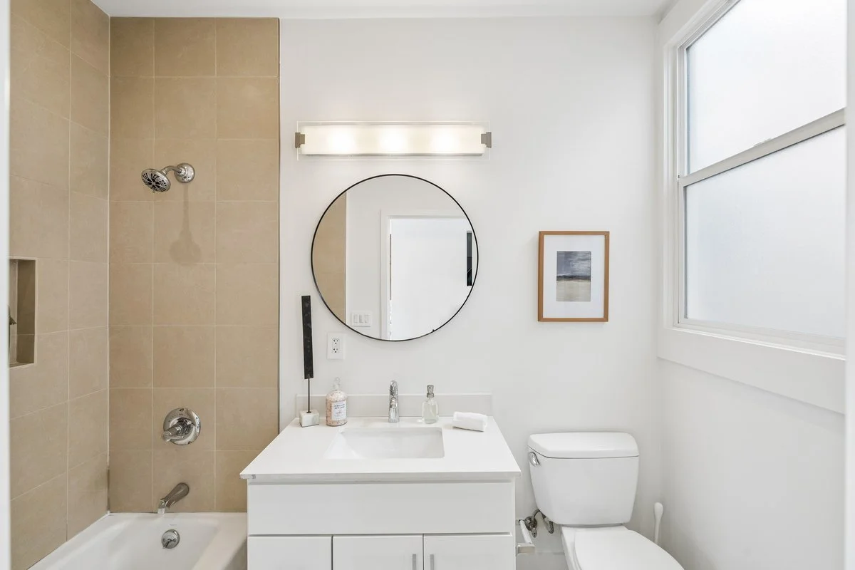 Modern bathroom with beige tiled shower area, white vanity with sink, round mirror, framed artwork, toilet, large window, and minimalistic decor.